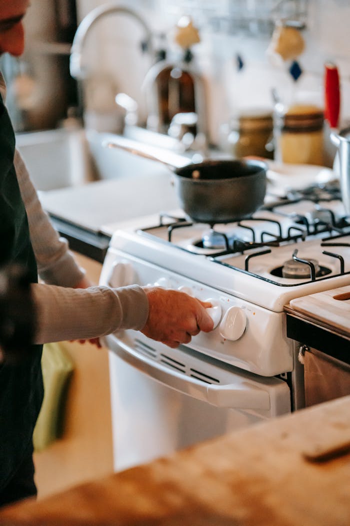 Crop anonymous male in casual clothes using gas stove with pan near counter and sink with tap near utensil in light kitchen at home